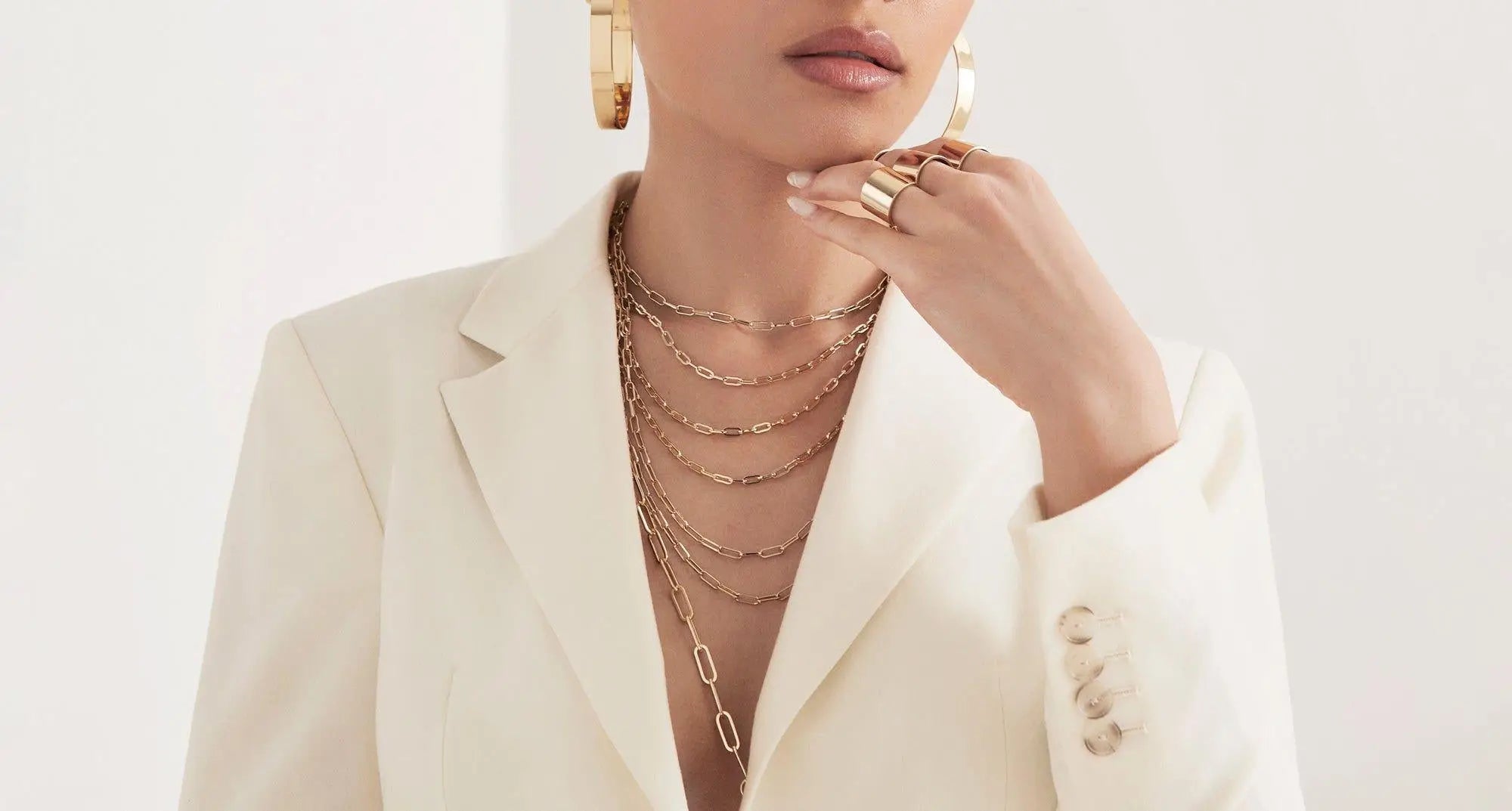 Woman in white blazer wearing layered gold necklaces hoop earrings and rings from MASK CHAINS collection