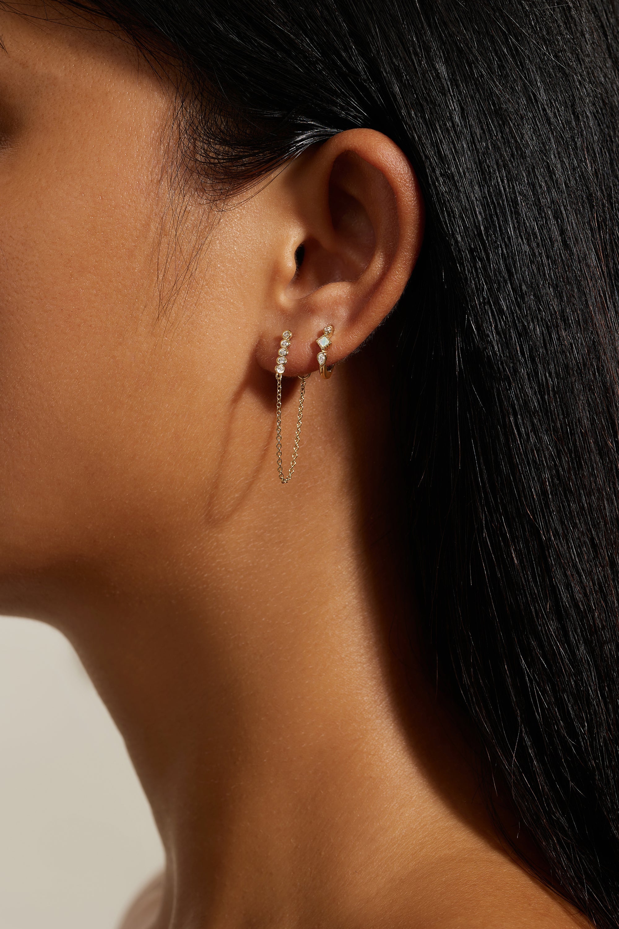 Woman’s ear with gold earrings including chain-connected studs and a single stud from Multi Shaped Opal and CZ Stone Huggie Hoops