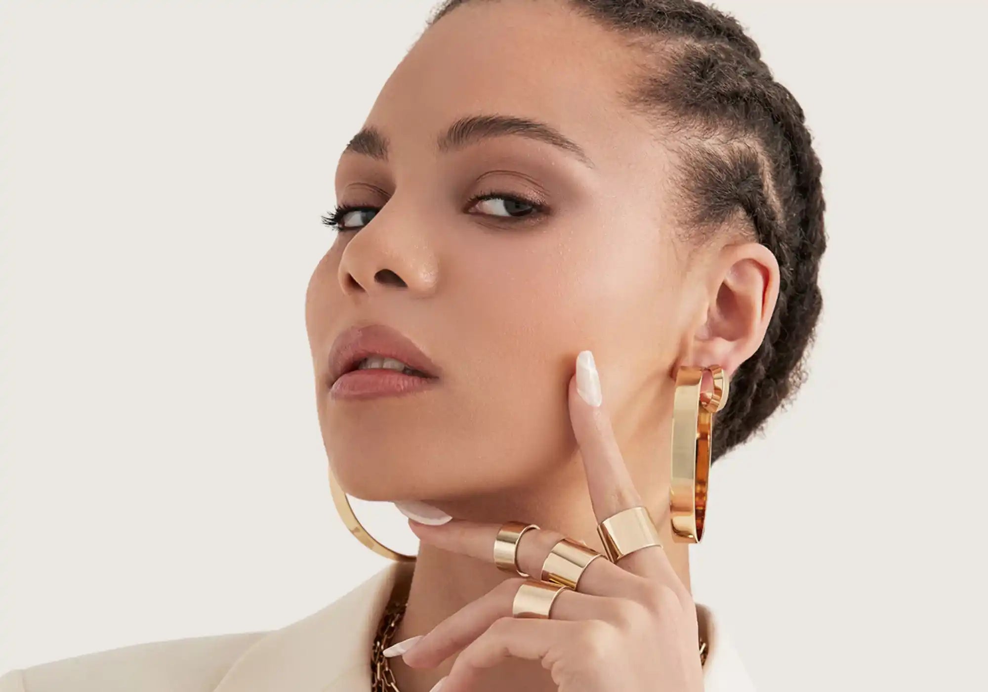 A woman with braided hair and light makeup, adorned with various gold jewelry including hoop earrings, a necklace, and multiple rings.