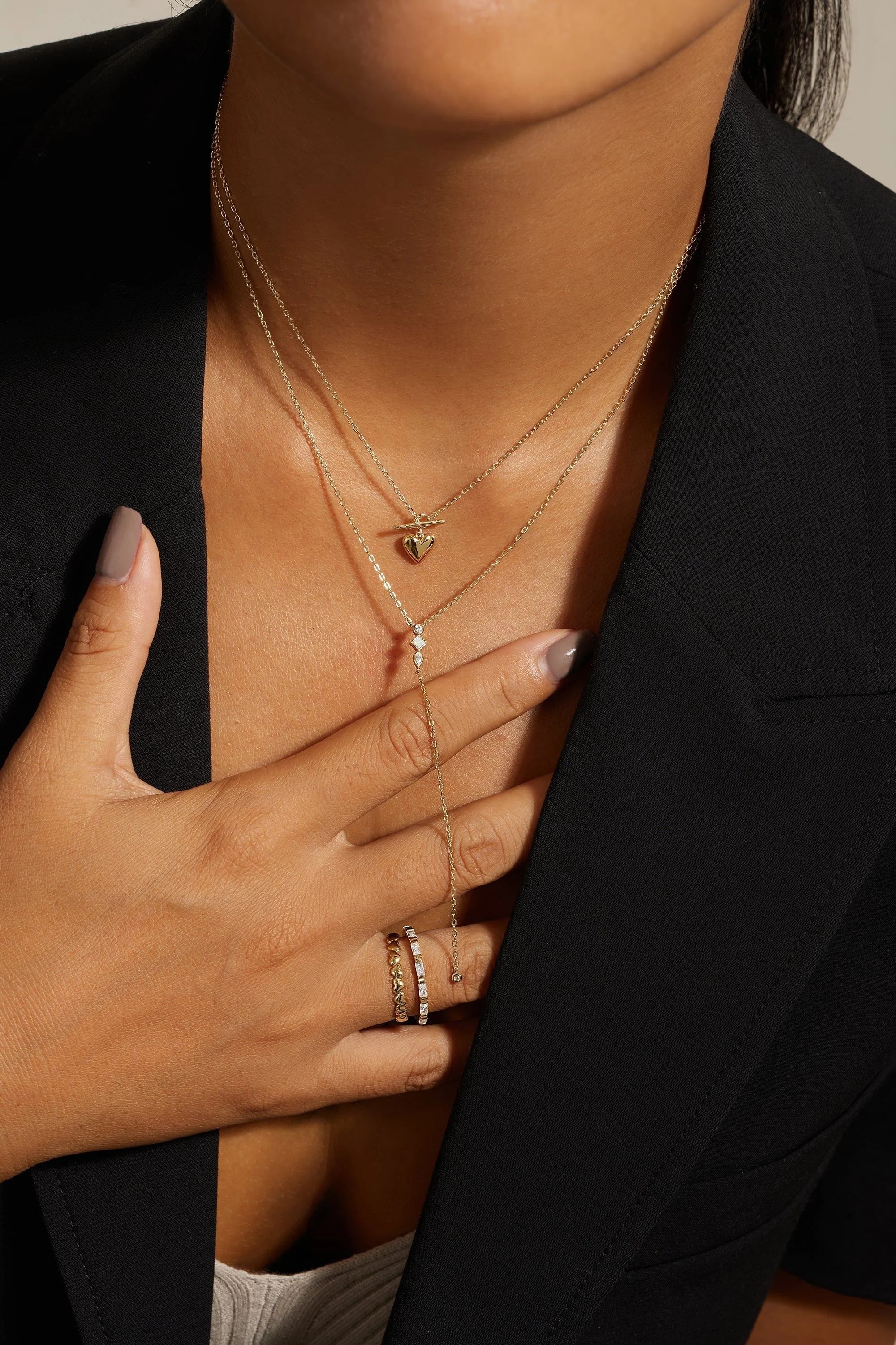 Woman wearing layered gold necklaces and rings with a dark blazer showcasing the Micro Heart Toggle Pendant Necklace